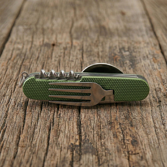 Close-up of a green 6-in-1 detachable camping multi-tool showing the integrated fork and corkscrew on a rustic wooden table.