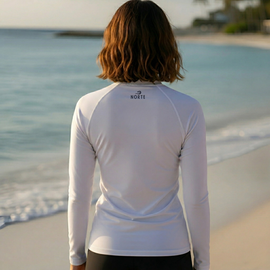 Rear view of a woman wearing the Norte Women's white long-sleeve rash guard on a beach, highlighting the small logo on the neckline and the comfortable, sun-safe performance fit.
