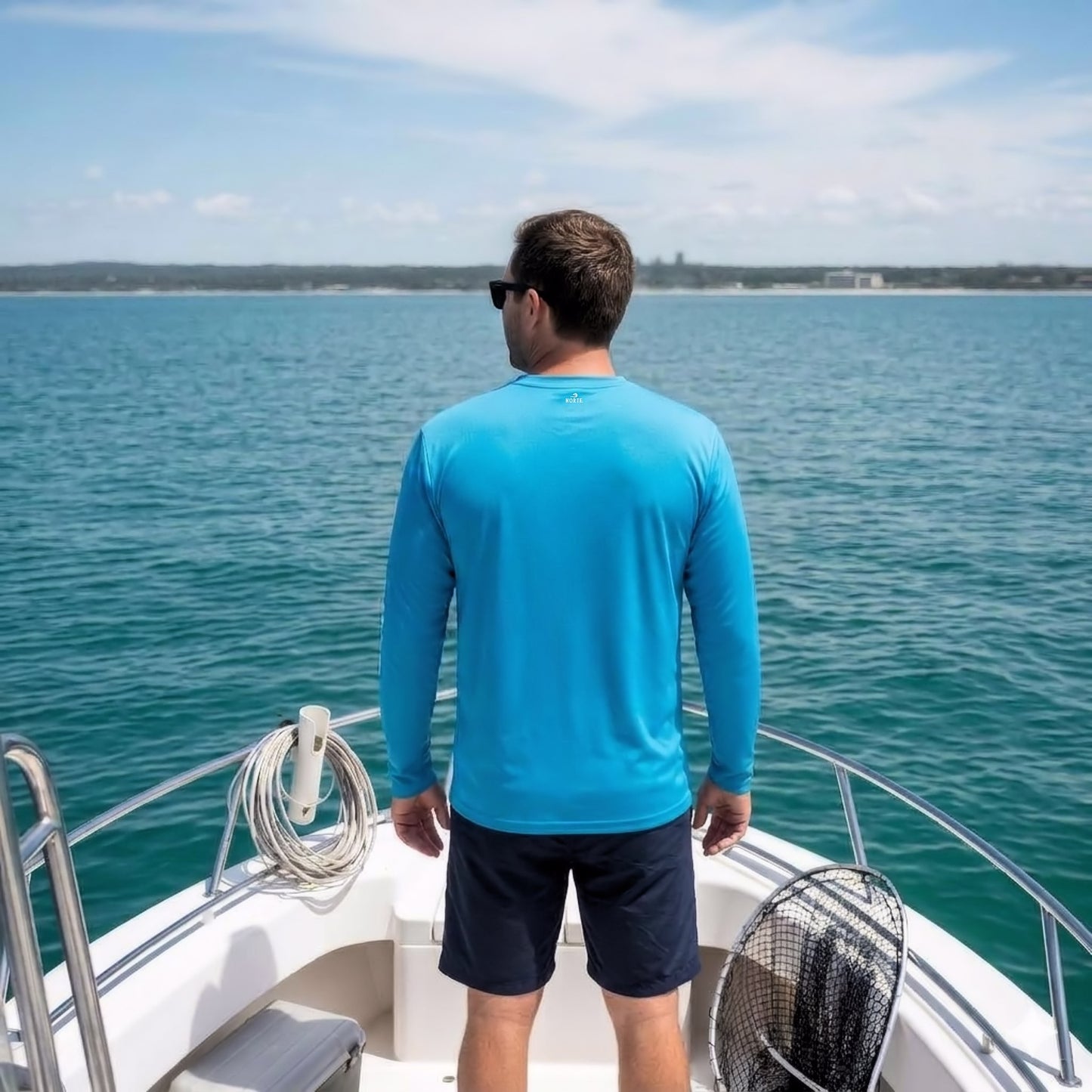 A man standing on the deck of a boat looking out at the water, wearing a blue long-sleeve performance fishing shirt and dark shorts.