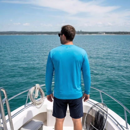 A man standing on the deck of a boat looking out at the water, wearing a blue long-sleeve performance fishing shirt and dark shorts.