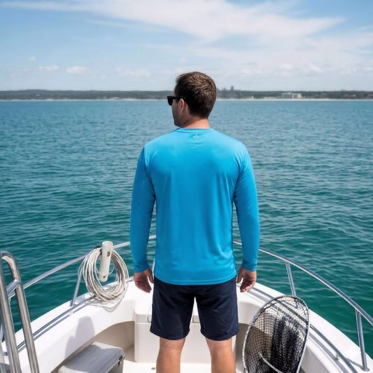 A man standing on the deck of a boat looking out at the water, wearing a blue long-sleeve performance fishing shirt and dark shorts.