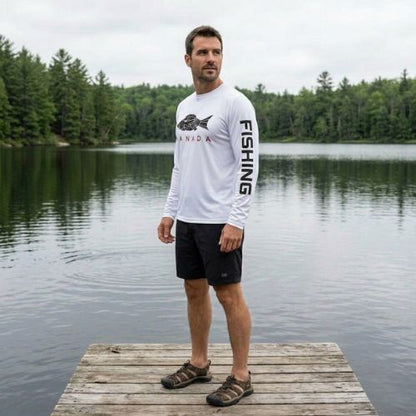 A side profile view of a man standing on a wooden dock by a calm lake with dense evergreen trees across the water. He wears a white long-sleeve fishing shirt showing a black fish graphic with "CANADA" on the chest and "FISHING" printed down the sleeve, styled with black shorts and dark hiking sandals.