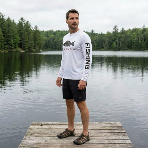 A side profile view of a man standing on a wooden dock by a calm lake with dense evergreen trees across the water. He wears a white long-sleeve fishing shirt showing a black fish graphic with "CANADA" on the chest and "FISHING" printed down the sleeve, styled with black shorts and dark hiking sandals.
