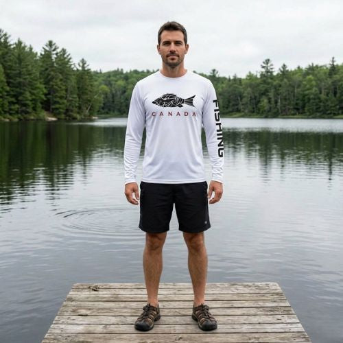 A man stands on a wooden dock by a calm lake with a background of evergreen trees. He is wearing a white long-sleeve performance shirt featuring a black fish graphic with the word "CANADA" and "FISHING" printed on the sleeve, paired with black athletic shorts and dark hiking shoes.