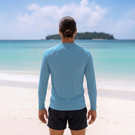 Back view of a man wearing a light blue long-sleeve relaxed fit rash guard and black boardshorts on a tropical beach background.