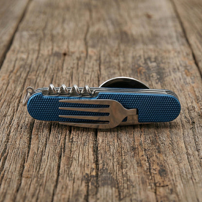 Close-up of a blue 6-in-1 detachable camping multi-tool showing the integrated stainless steel fork and corkscrew features on a rustic wooden table.