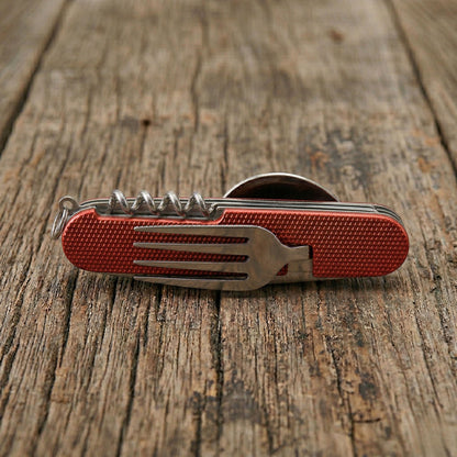 Close-up of a red 6-in-1 detachable camping multi-tool showing the integrated stainless steel fork and corkscrew on a rustic wooden table.
