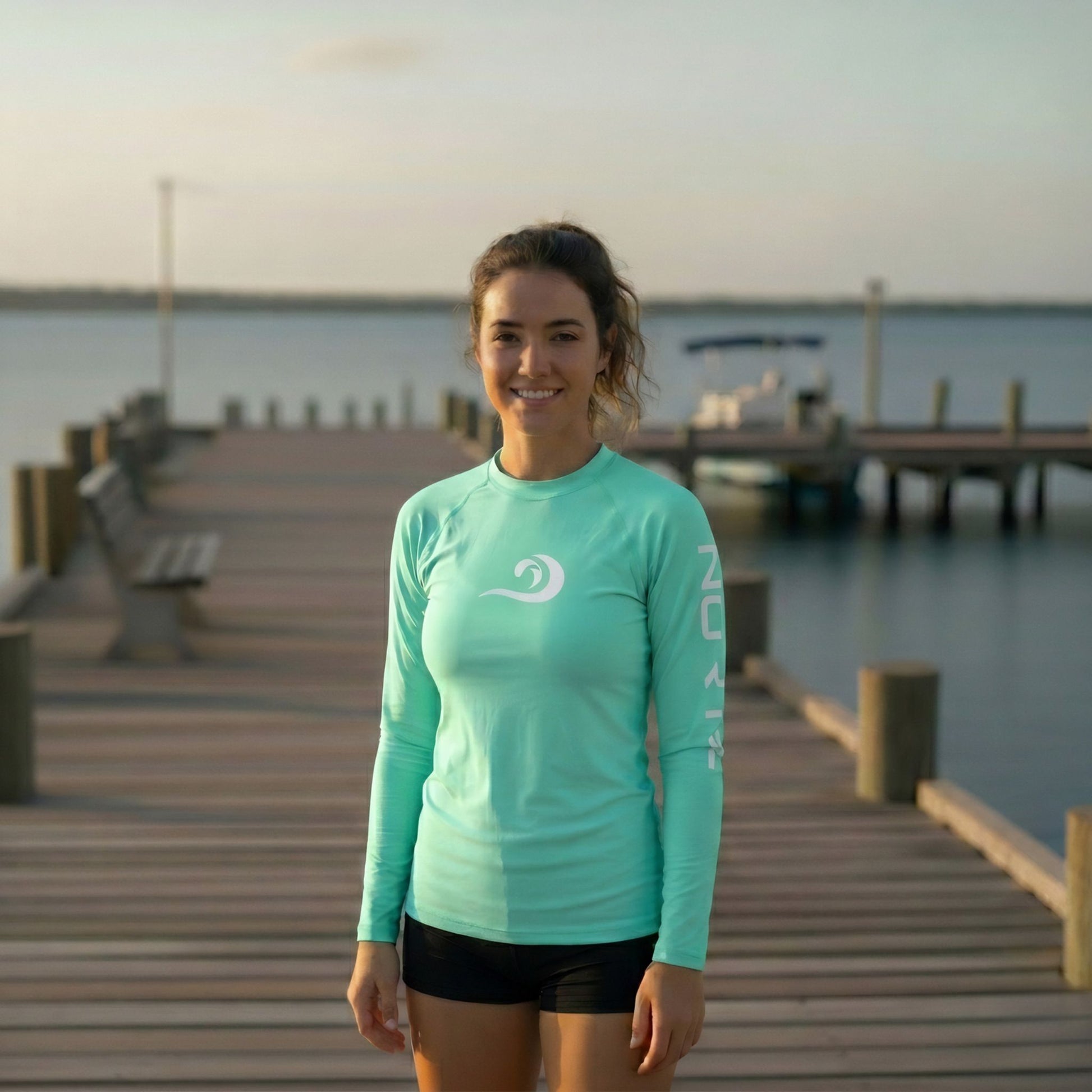 Woman wearing the Norte Women’s Performance Rash Guard in Mint on a dock at sunset, featuring the white wave-mark logo and UPF 50+ sun protection.