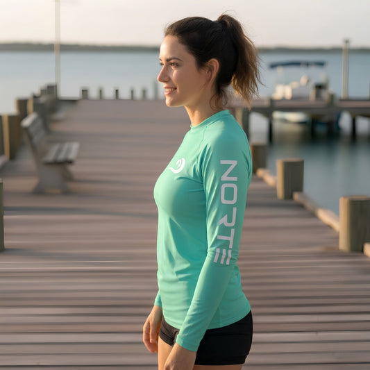 A woman standing in profile on a wooden dock by the water, wearing a Norte long-sleeve mint green rash guard with a white logo on the chest.