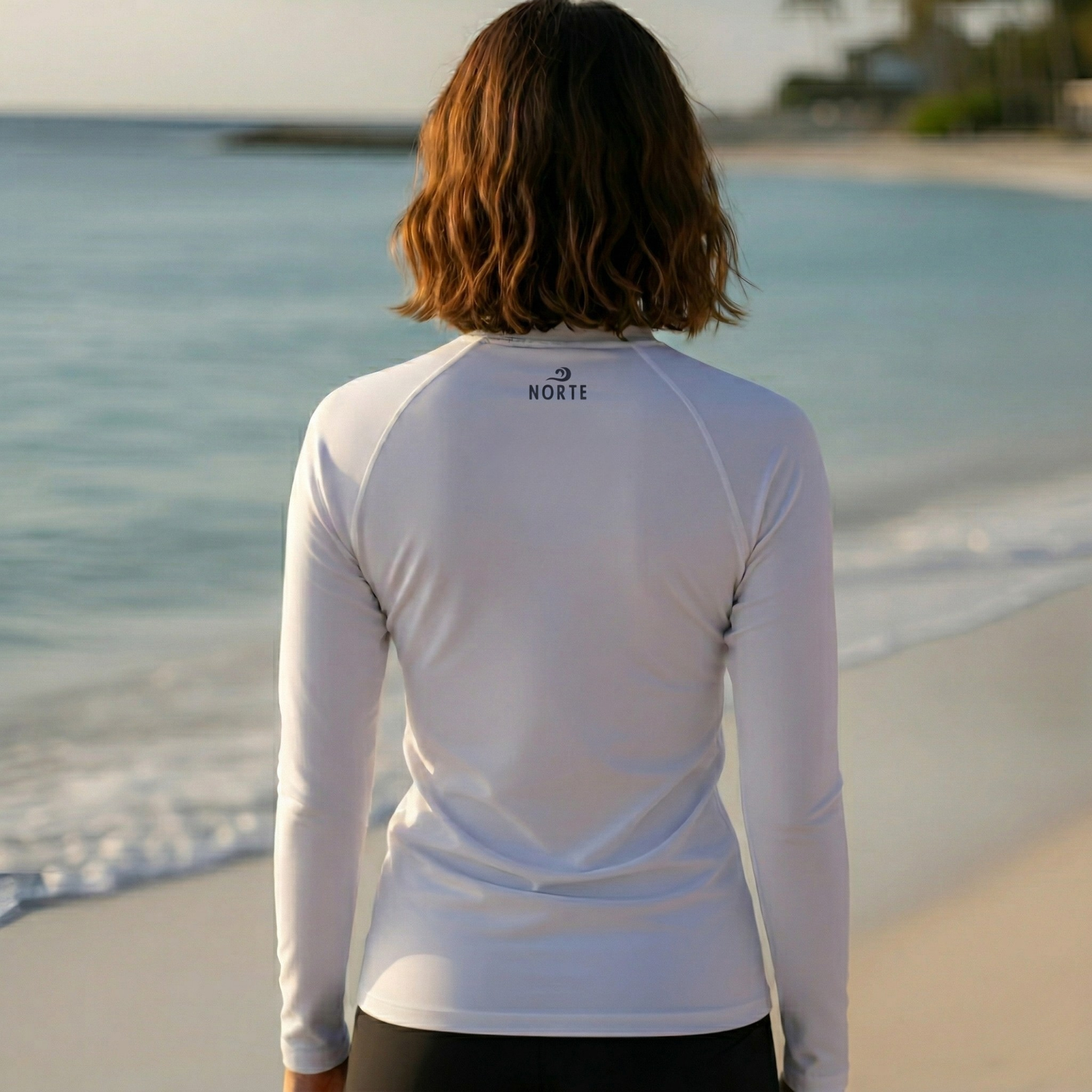 Rear view of a woman wearing the Norte Women's white long-sleeve rash guard on a beach, highlighting the small logo on the neckline and the comfortable, sun-safe performance fit.